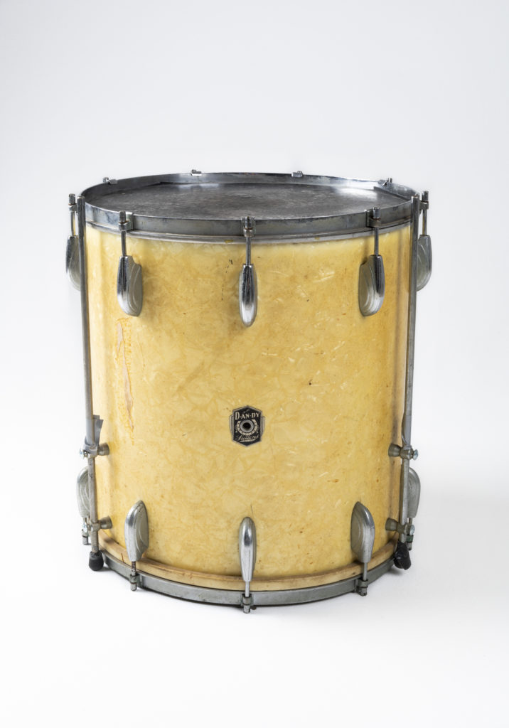 Floor-tom featuring a pearl celluloid laminate over the plywood shell. There are chrome-plated fittings running around the circumference of the drum at the top and bottom. A badge features the Dandy logo with the words ‘Supplied by Suttons Pty Ltd’.
